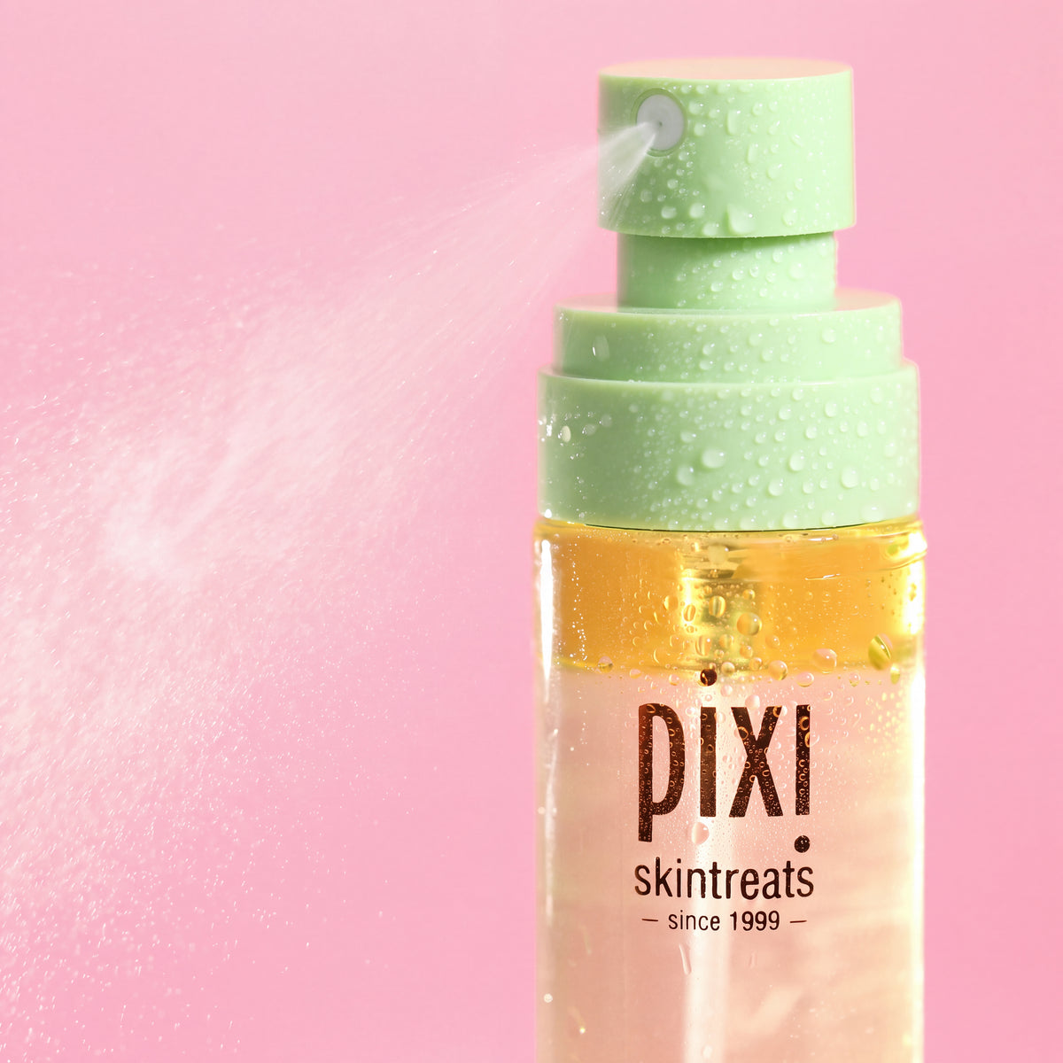 Pixi Skintreats facial mist bottle with a light green pump dispenser spraying a fine mist, set against a solid pink background.